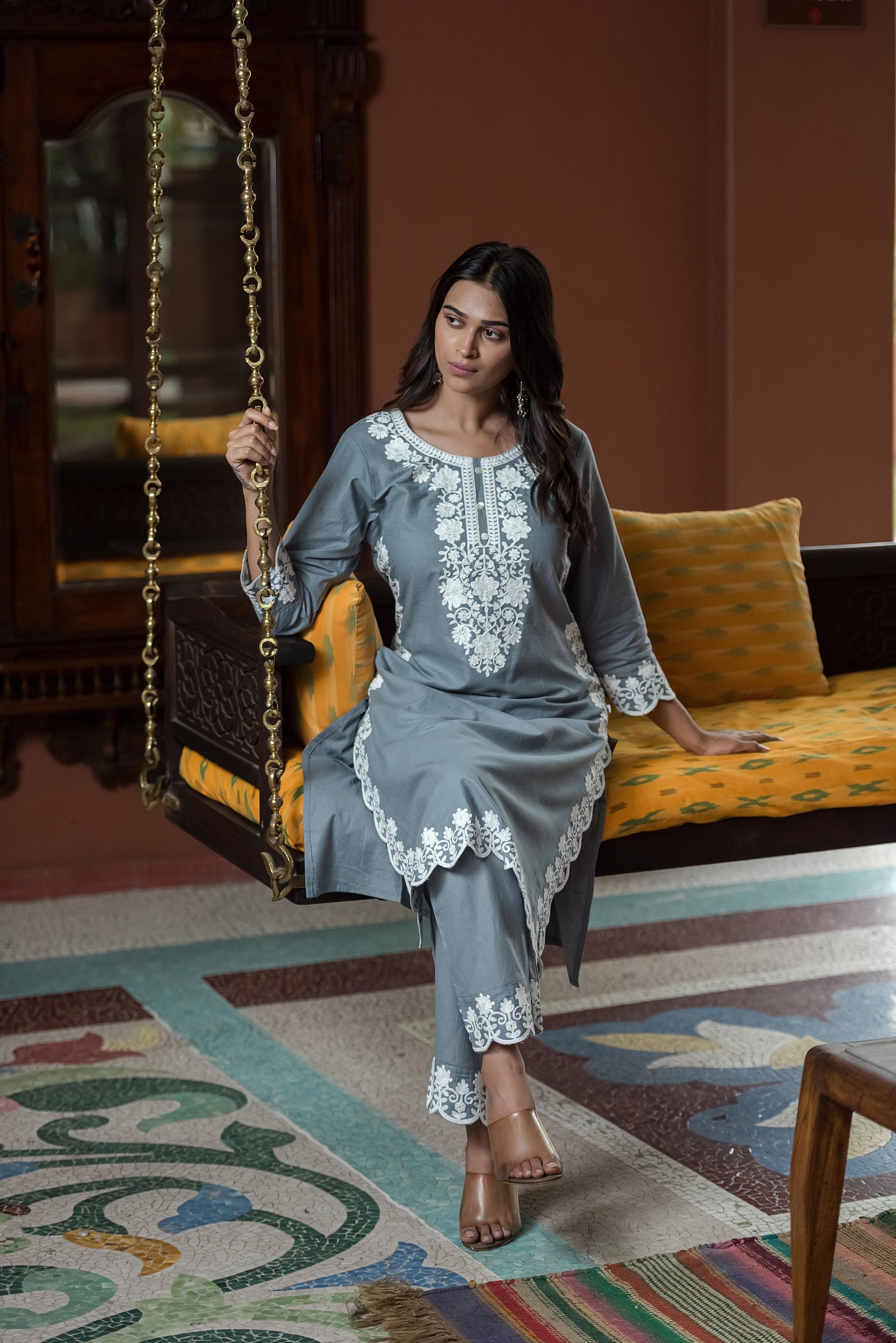 Woman in a gray embroidered outfit sitting on a colorful rug with a swing in the background.